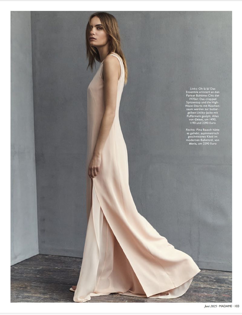 Fashion model wearing a light pink gown with a slit poses in a studio against a gray background. Full body shot.