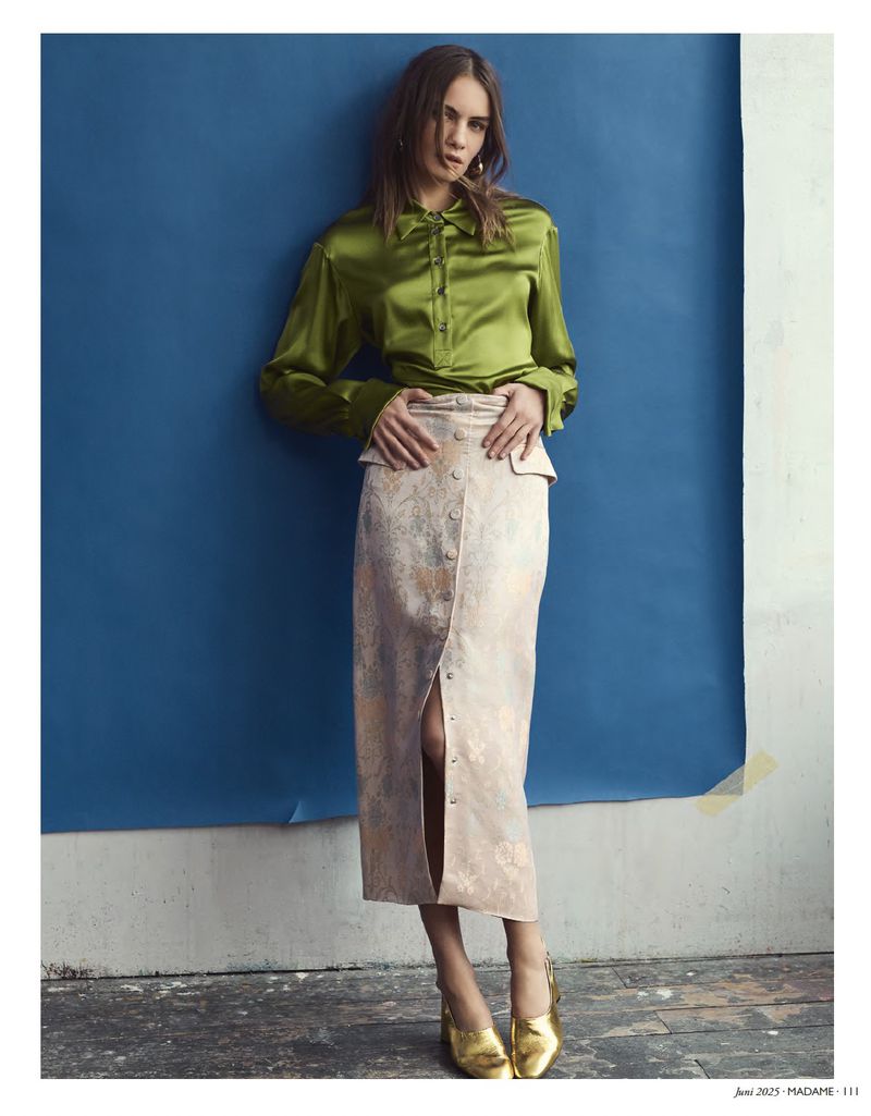 A woman models a shiny green collared long-sleeve shirt and patterned beige skirt against a blue backdrop in a fashion editorial shot.
