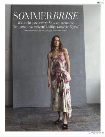 Fashion editorial shot of a woman in a floral dress standing indoors against a gray backdrop, paired with black sandals in a full shot.
