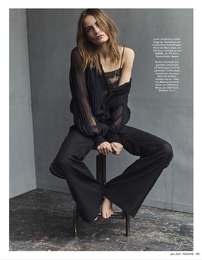 Editorial fashion portrait of a woman in black lingerie and sheer blouse, seated on a stool against a grey background.