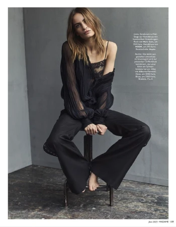 Editorial fashion portrait of a woman in black lingerie and sheer blouse, seated on a stool against a grey background.