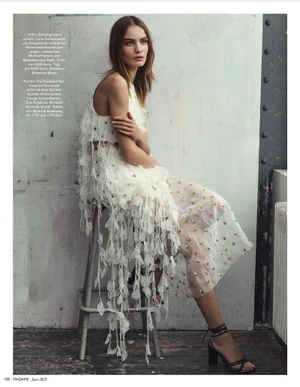 Model seated on a metal stool wearing a white sheer embroidered dress against a textured industrial wall
