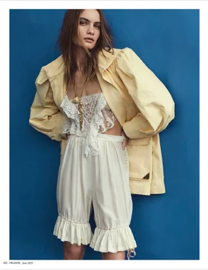 Model in yellow jacket and white lace-trimmed romper against a solid blue background for Madame Germany editorial.