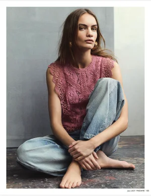 Model Nina Marker seated on the floor wearing a pink lace top and light blue denim jeans against a grey studio backdrop.