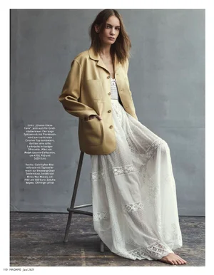 Model Nina Marker seated on a stool wearing a yellow leather jacket and white lace dress against a grey studio wall