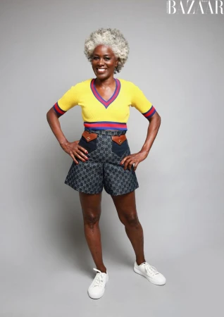 A full-length portrait of a mature Black woman with gray hair in a yellow top and Gucci shorts, posing against a neutral gray backdrop.
