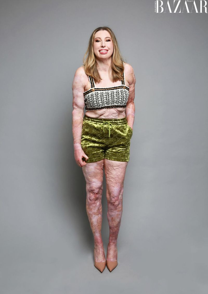 Full-length shot of an adult woman posing in a studio, wearing a knit crop top, green velvet shorts, and nude heels against a gray backdrop.