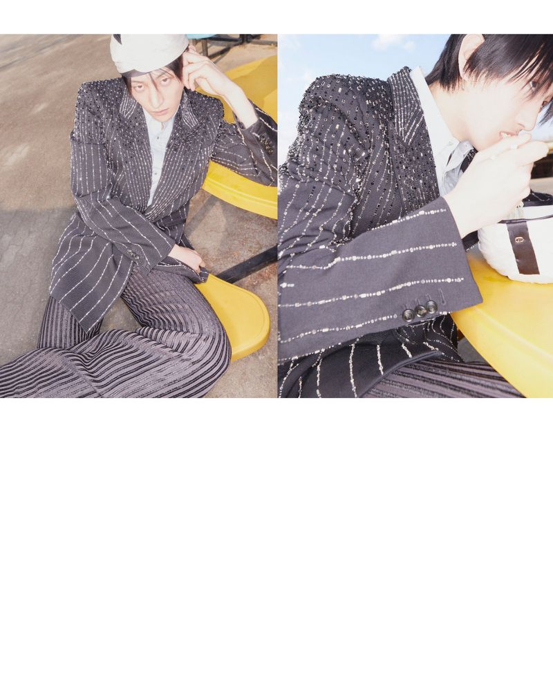 Editorial fashion shot: androgynous model in a striped suit with a white cap, sitting on a yellow chair in an indoor setting.