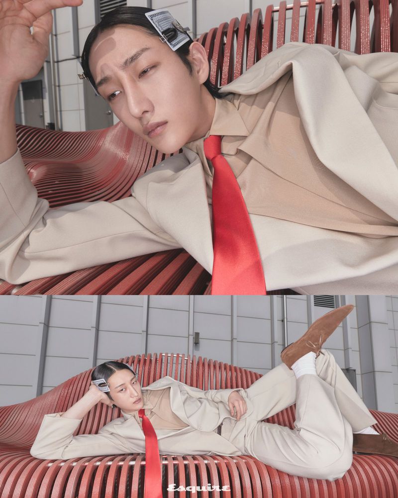 A stylish Asian male model, with a serious expression, poses on a red slat bench in a tan suit with a red tie for an editorial fashion shot.