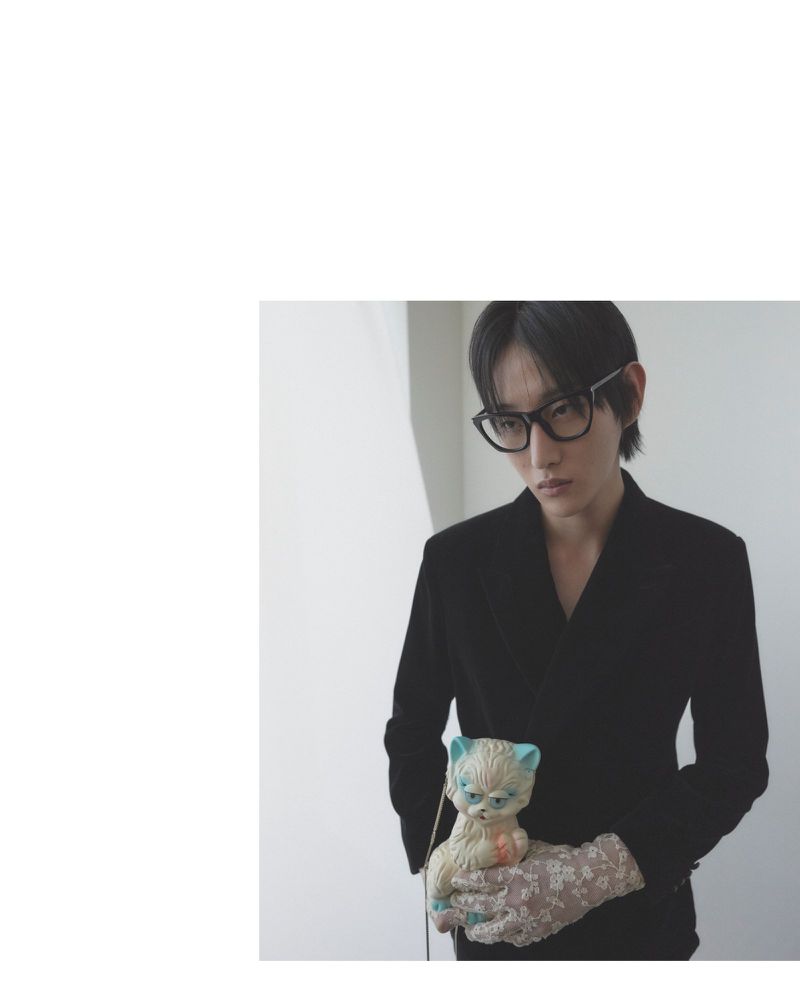 Adult male with dark hair and glasses in a black jacket holding a vintage cat doll with patterned gloves indoors near a white wall.
