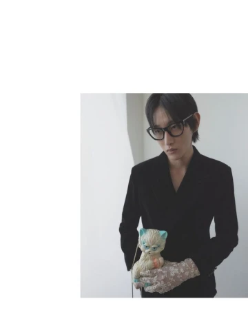 Adult male with dark hair and glasses in a black jacket holding a vintage cat doll with patterned gloves indoors near a white wall.