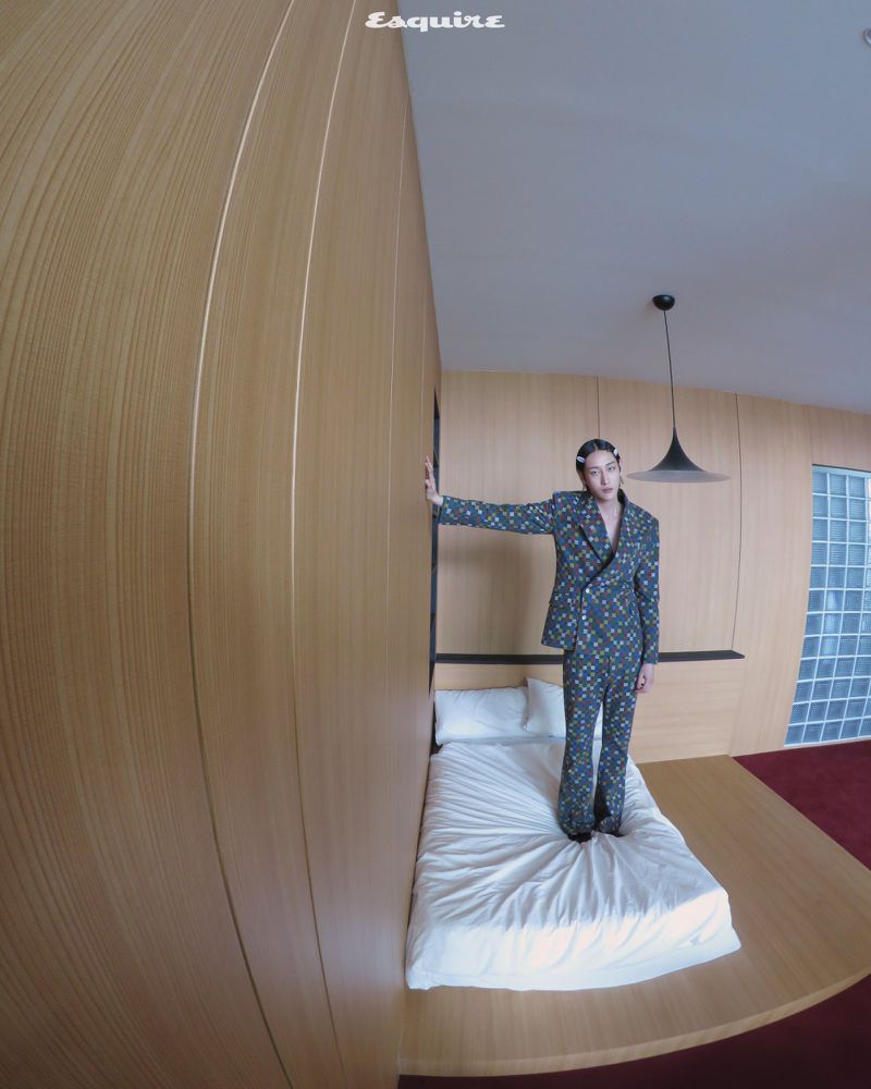 Adult male in a patterned suit stands on a bed in a studio with light wood walls and red carpet, lit by an overhead pendant lamp.