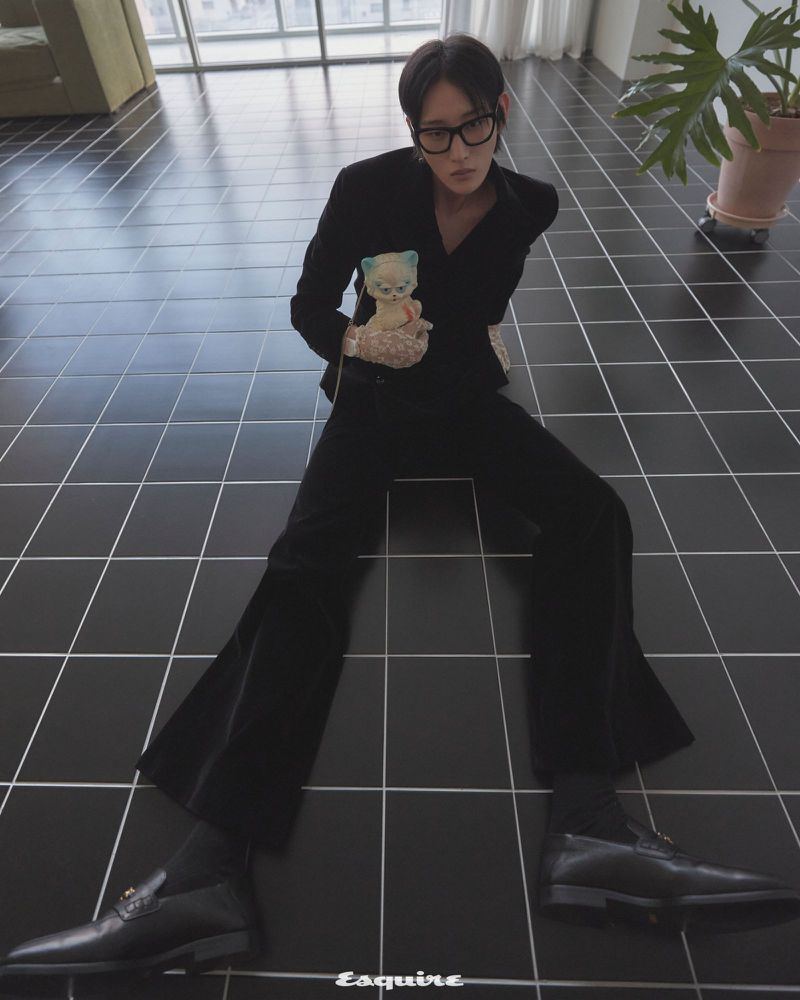 A person in a black suit and glasses sits on a black tiled floor, holding a doll, with a potted plant and window in the background.