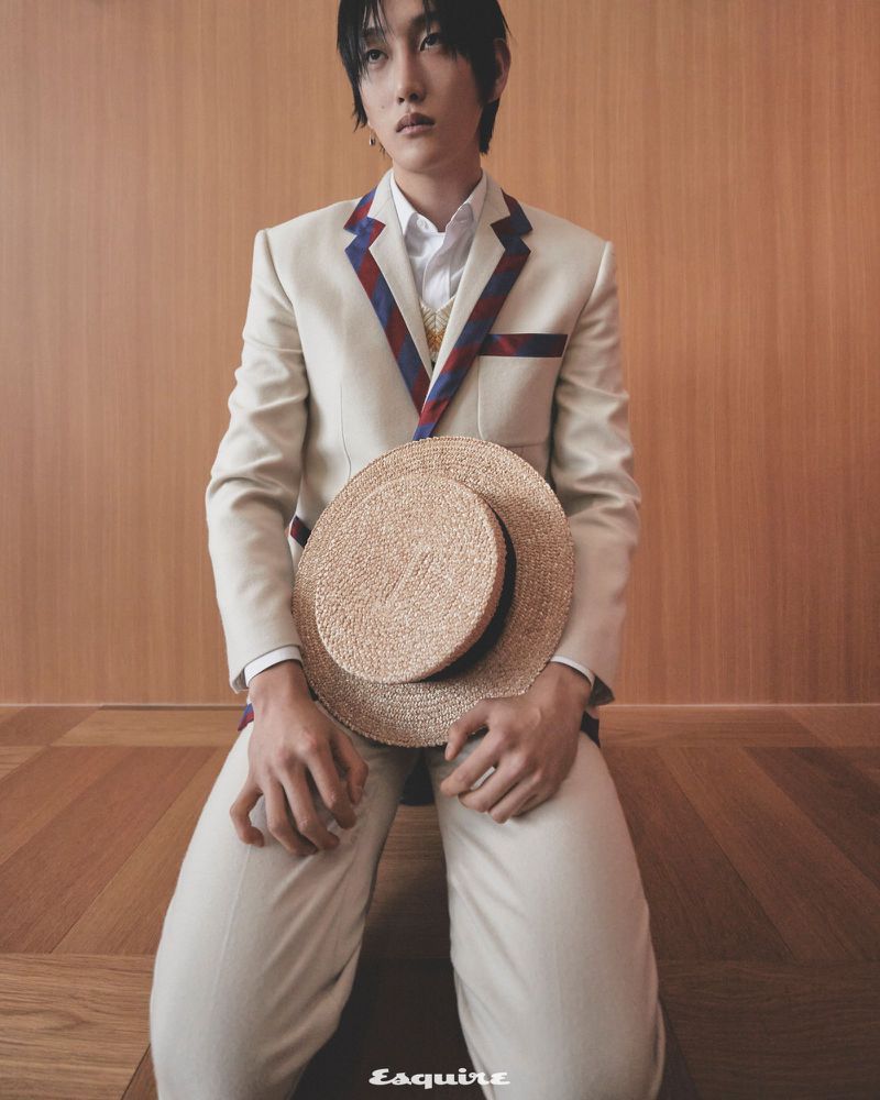 Portrait of adult male kneeling indoors with straw hat in hands and wearing white jacket and pants and striped shirt collar.