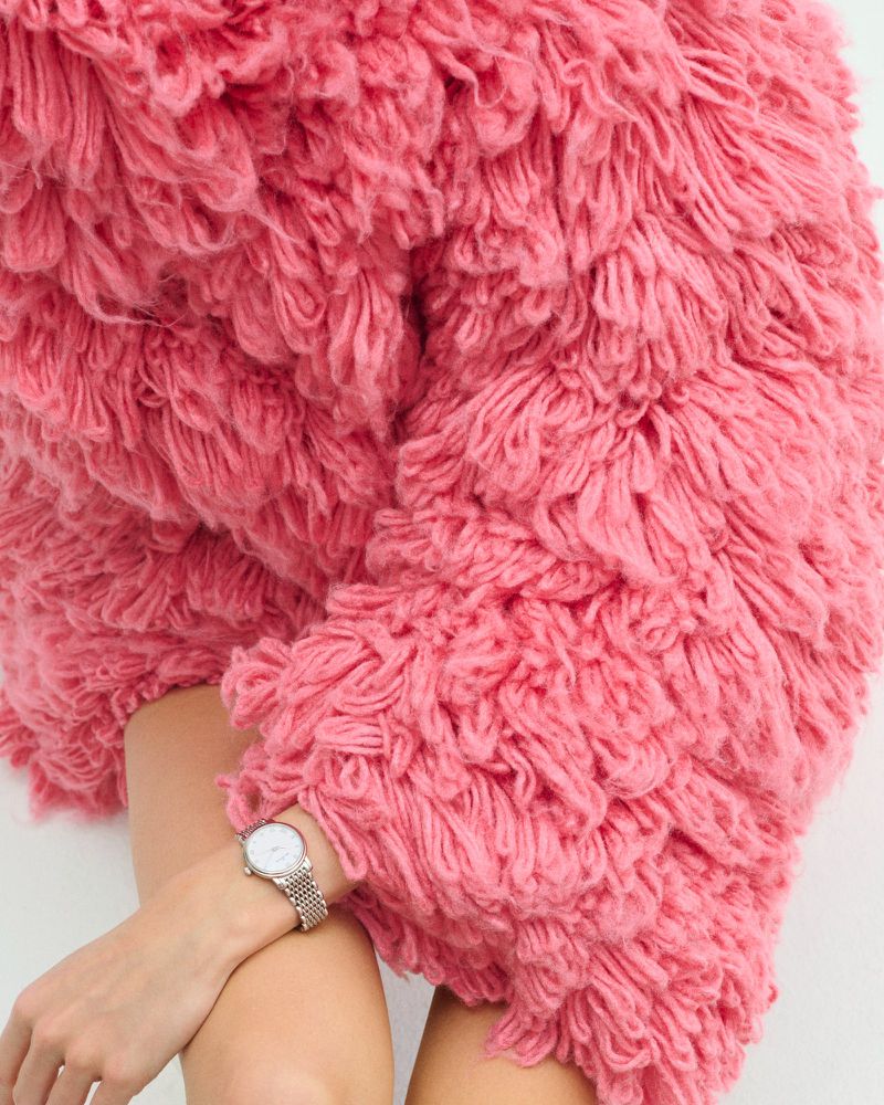 Woman wearing pink shaggy textured coat with white dial watch and silver mesh strap against a white backdrop, fashion still life.