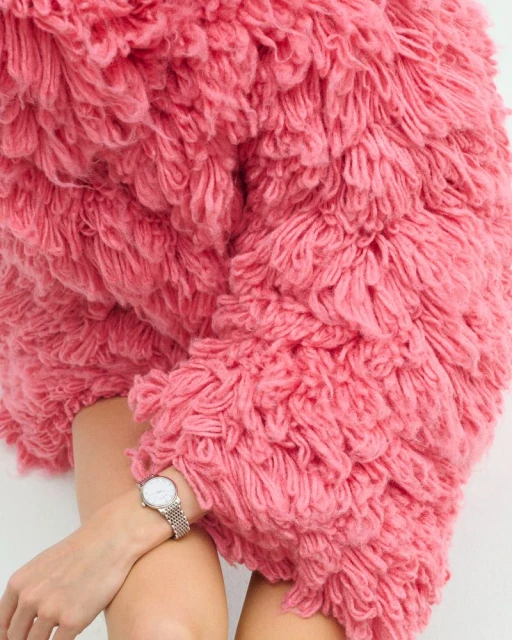 Woman wearing pink shaggy textured coat with white dial watch and silver mesh strap against a white backdrop, fashion still life.