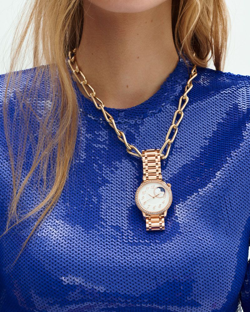 A woman wears a gold chain necklace with a rose gold wristwatch as a pendant, complemented by a blue sequined top in a studio setting.