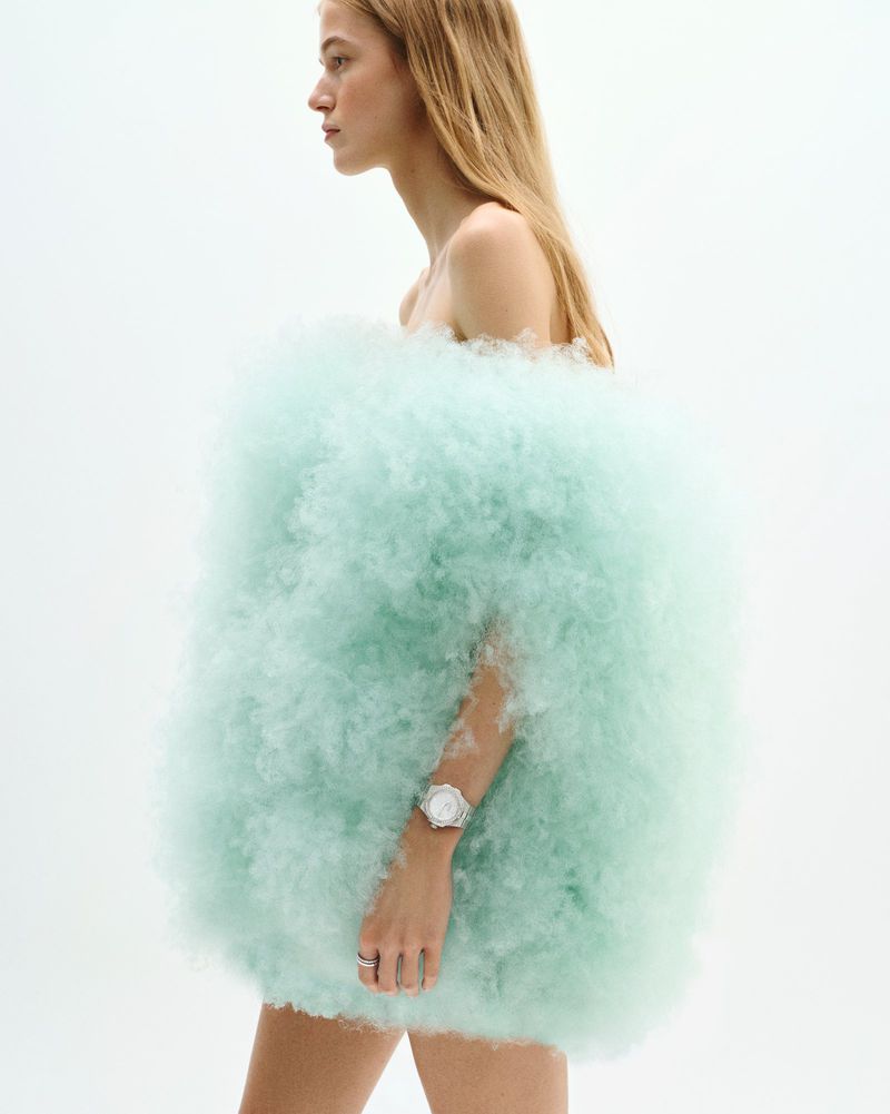 A blonde woman with a white watch poses in front of a white background, wearing a light-green cloud-like fluffy material as clothing.