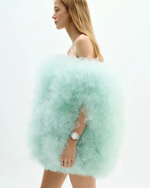 A blonde woman with a white watch poses in front of a white background, wearing a light-green cloud-like fluffy material as clothing.
