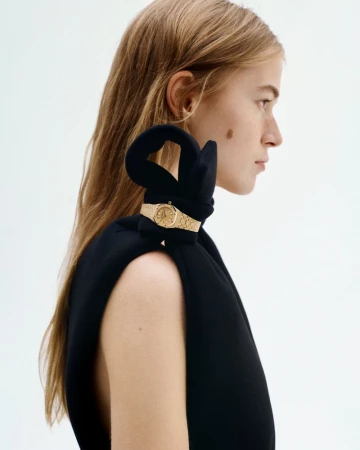 Profile of a blonde woman in a black top with a wristwatch on her shoulder against a stark white background.