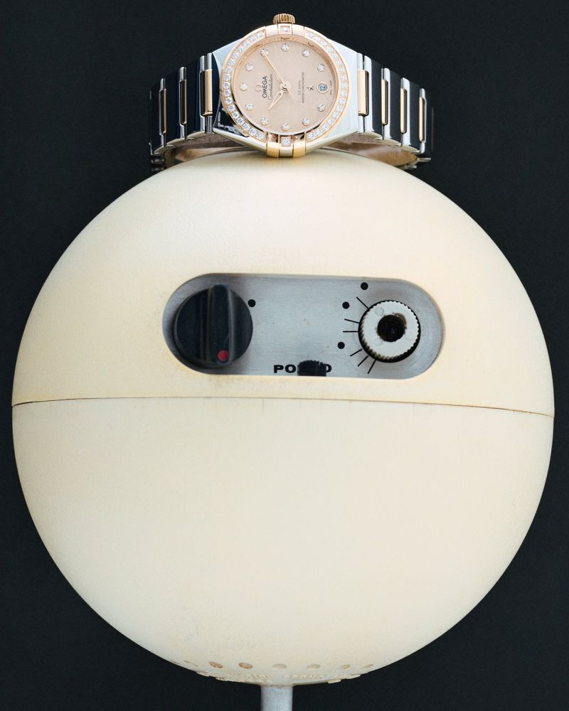 Wristwatch with a diamond bezel is placed atop a vintage spherical speaker with black knob details on a black background.