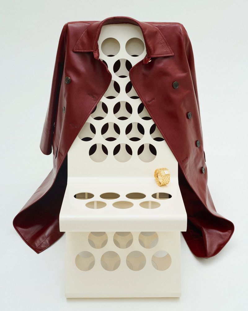 Maroon leather trench coat draped over a modern off-white display stand with circular cutouts and a golden ring on the top shelf.
