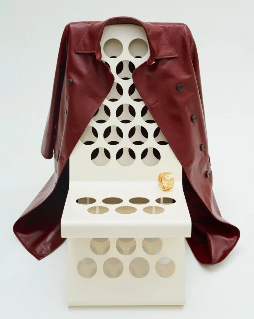 Maroon leather trench coat draped over a modern off-white display stand with circular cutouts and a golden ring on the top shelf.