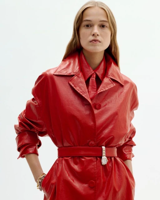 Blonde female poses in shiny red trench coat with matching collared shirt and tie against a bright white seamless backdrop.