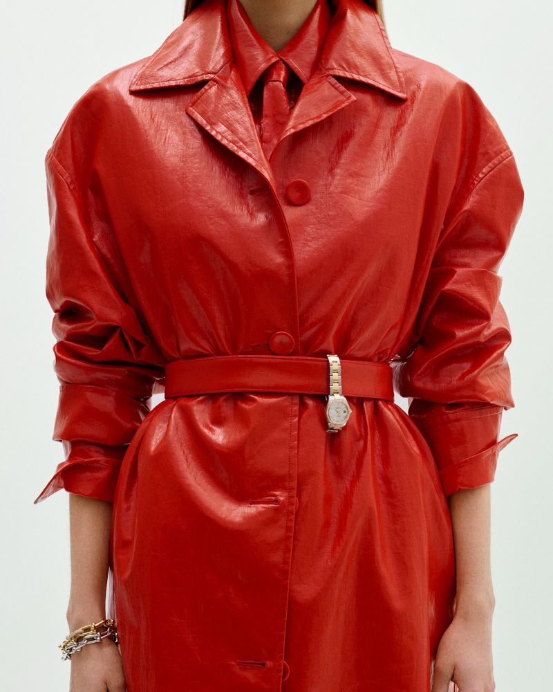 Close-up of a person in a shiny red trench coat with a matching shirt and tie, accessorized with a belt, wristwatch, and bracelets.