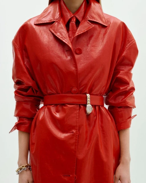 Close-up of a person in a shiny red trench coat with a matching shirt and tie, accessorized with a belt, wristwatch, and bracelets.