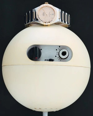 Omega luxury wristwatch with diamond bezel resting atop a spherical cream-colored vintage device against a solid black background.