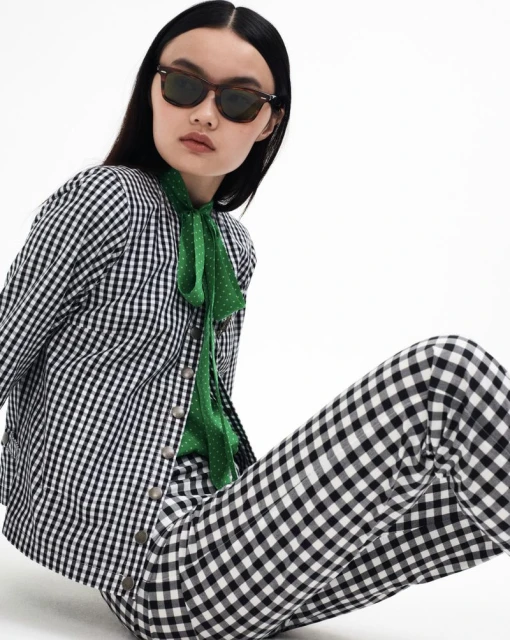 Young woman in black and white gingham jacket and pants poses on a white backdrop, wearing brown sunglasses and a green polka dot blouse.