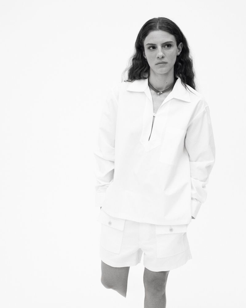 Black and white shot of a woman in a white collared shirt and shorts against a bright backdrop. She has her hands in her pockets.