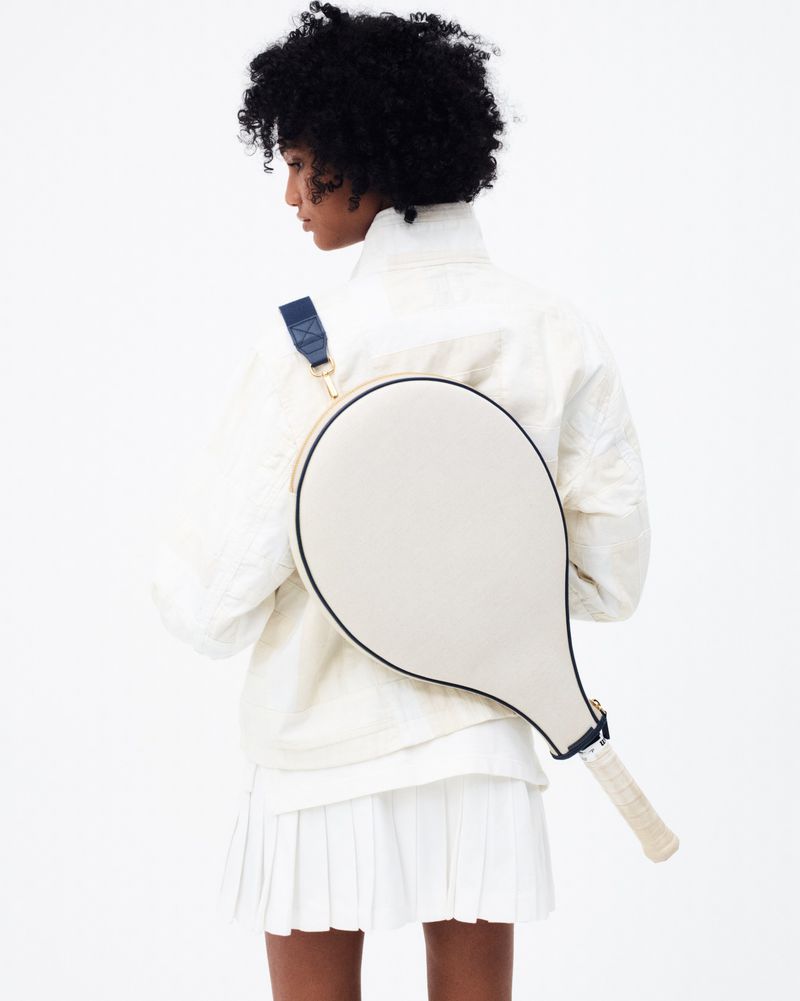 Back view of a dark-haired woman with a tennis racquet on her back wearing a white jacket and skirt, against a white backdrop.