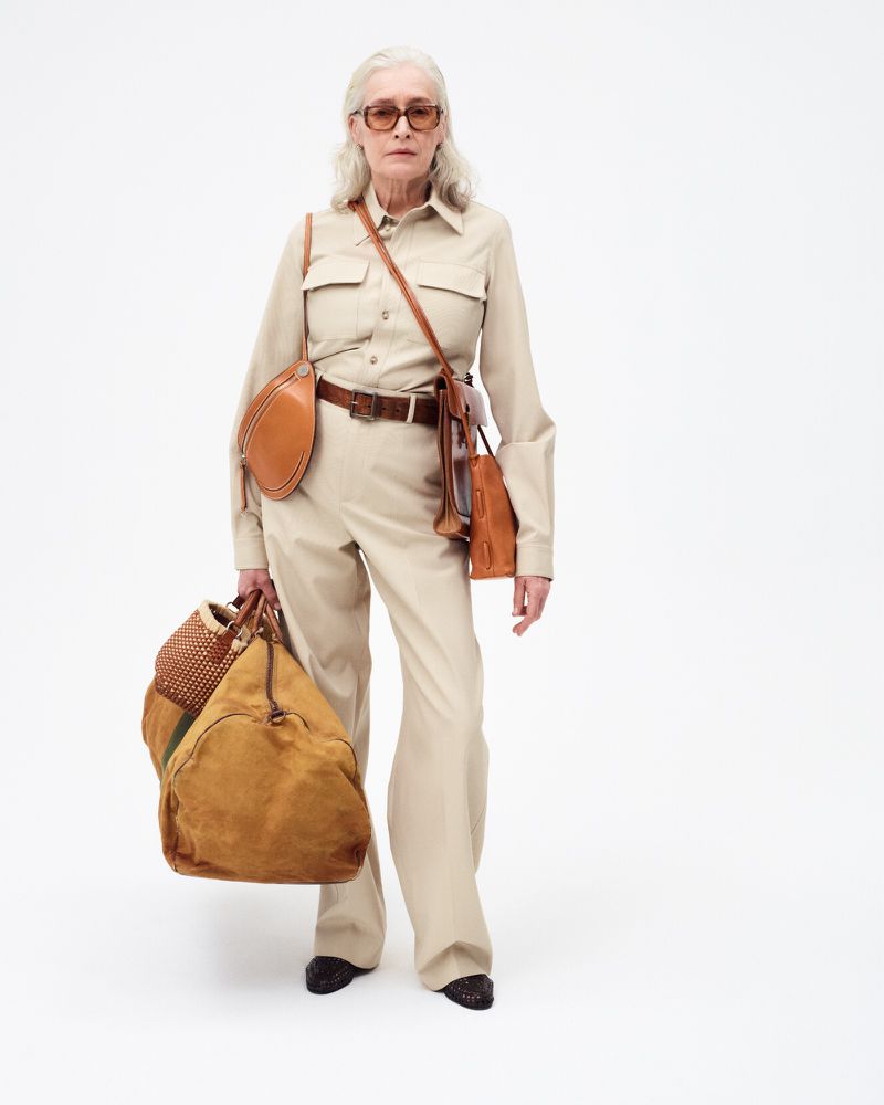Mature woman in tan outfit carrying brown leather handbags and suede duffel bag, standing in white studio. Fashion editorial shot.