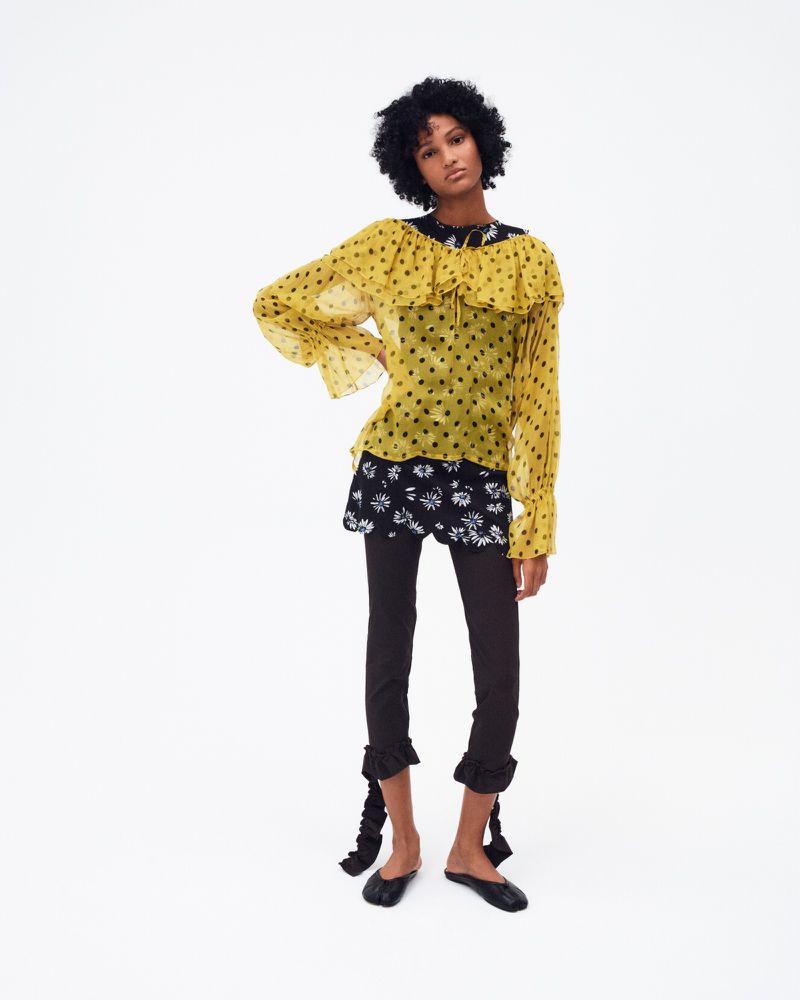 Fashion model poses in a yellow polka dot blouse, black floral shorts, and black leggings against a white backdrop in a studio shot.