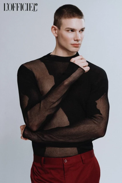 Portrait of a young adult male fashion model in a sheer black long-sleeved shirt and red trousers against a white background.