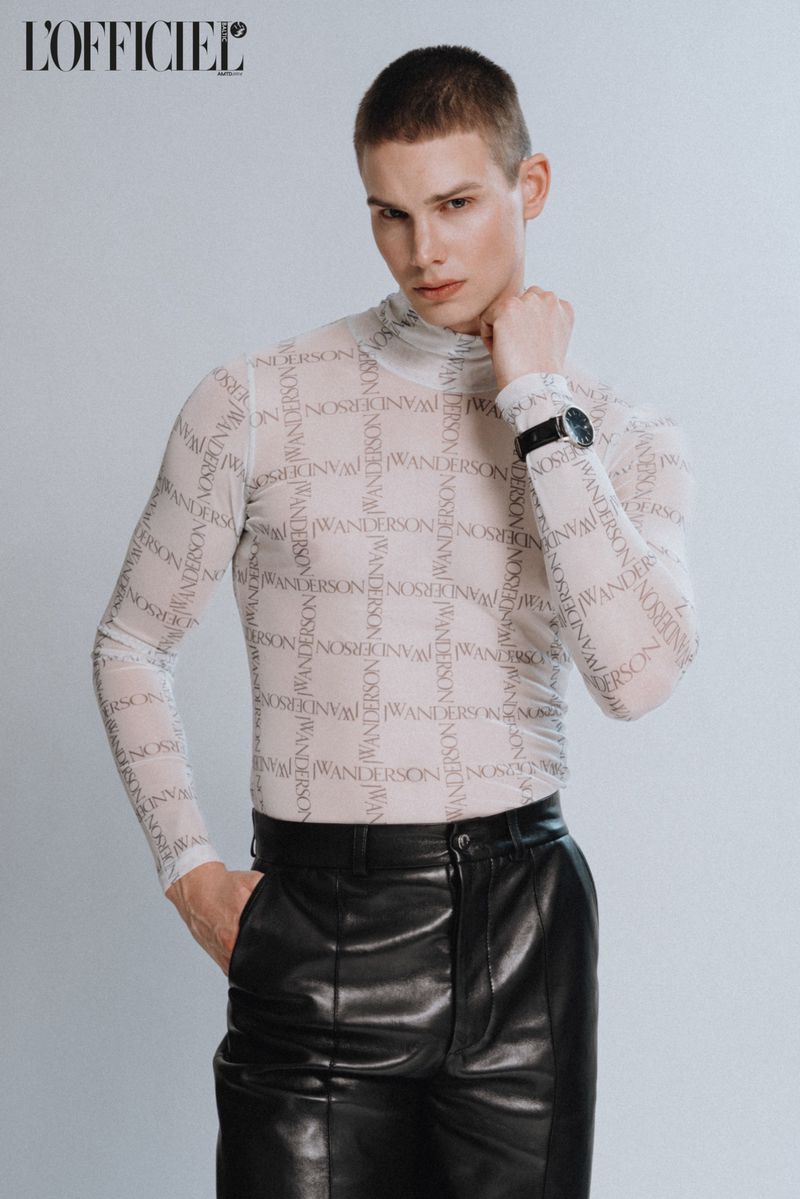 Headshot of a young adult male in a sheer logo-print shirt, dark pants, and a wristwatch, posing in a photo studio with neutral lighting.