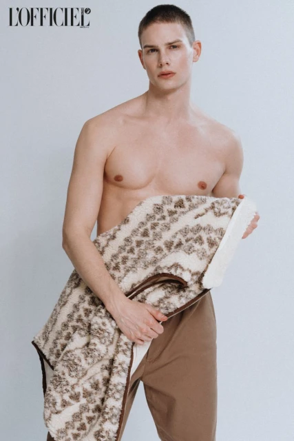Topless male fashion model poses indoors, holding a patterned blanket. He wears khaki pants against a white studio backdrop with magazine logo.