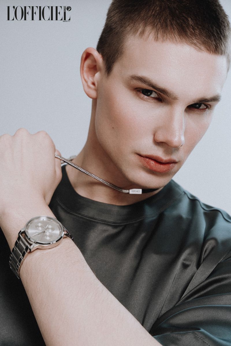 Close-up fashion shot of a young adult male model wearing a gray top, chain necklace, and wristwatch in a studio setting.