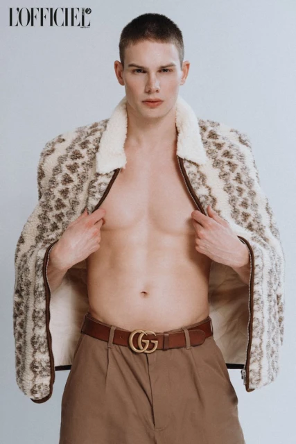 Fashion portrait of a young male model wearing a patterned jacket and brown pants with a 'G' logo belt in a studio setting.