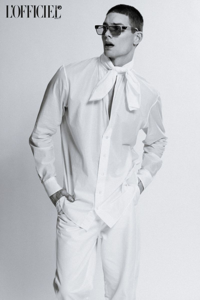 Black-and-white fashion shot of a male model wearing a white collared shirt, loose white pants, and sunglasses posing indoors against a solid backdrop.