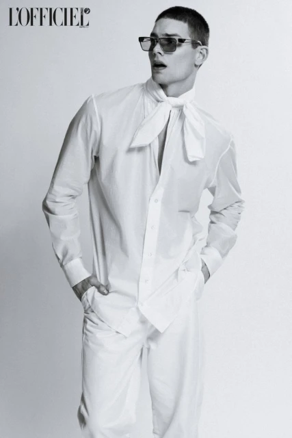 Black-and-white fashion shot of a male model wearing a white collared shirt, loose white pants, and sunglasses posing indoors against a solid backdrop.