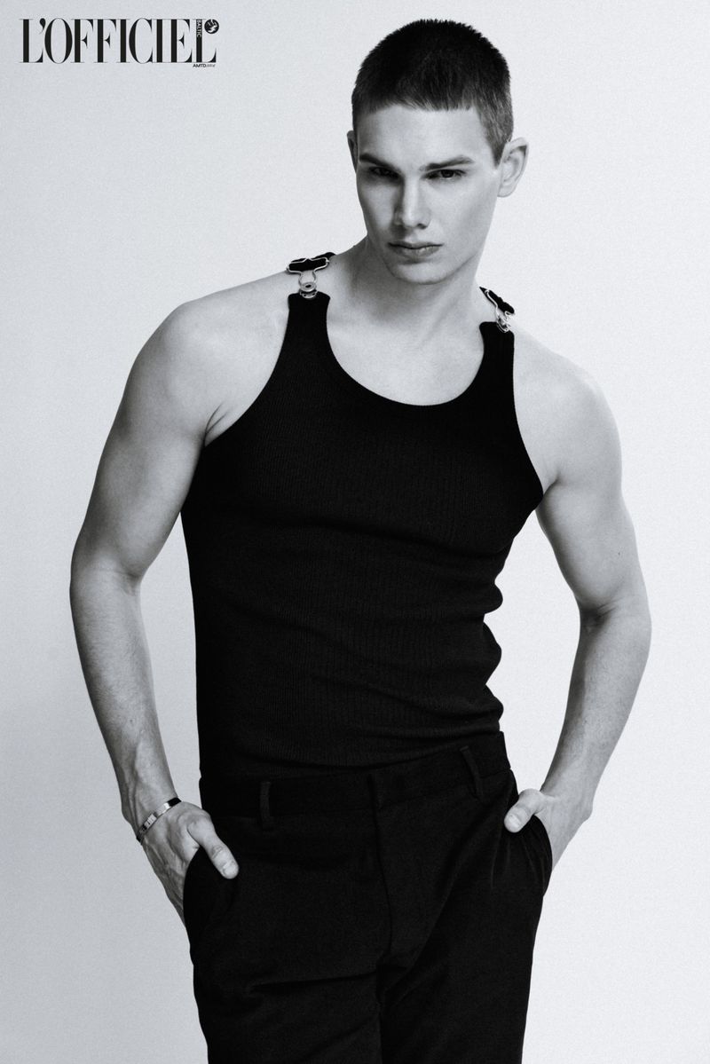 Black and white portrait of a young adult male model in a black tank top and pants, posing indoors with hands in pockets.