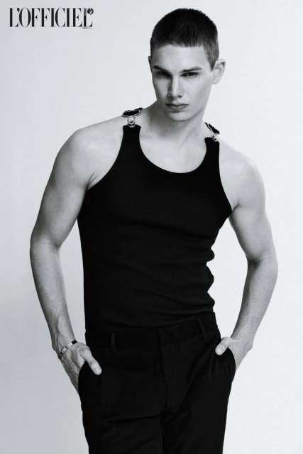 Black and white portrait of a young adult male model in a black tank top and pants, posing indoors with hands in pockets.