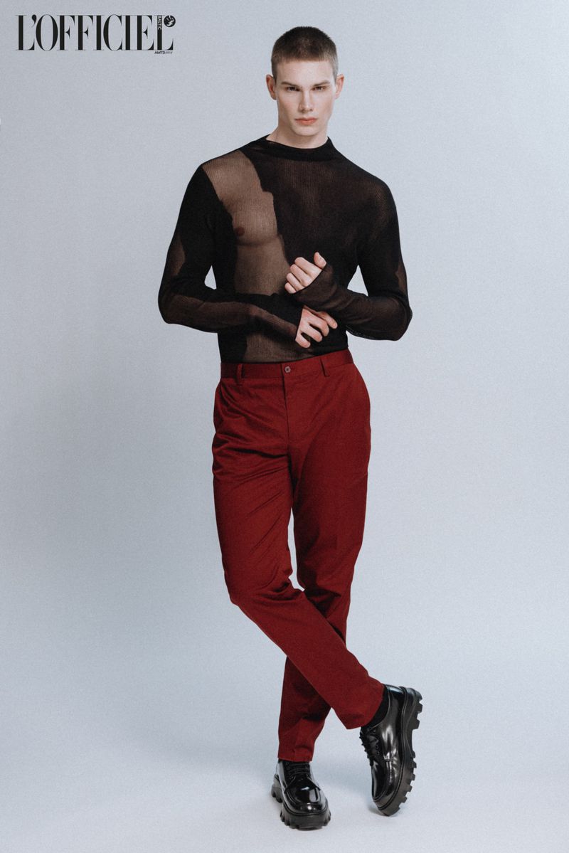 Male model posing in a studio wearing a black sheer top and burgundy pants, glossy black shoes, and a buzzcut hairstyle.