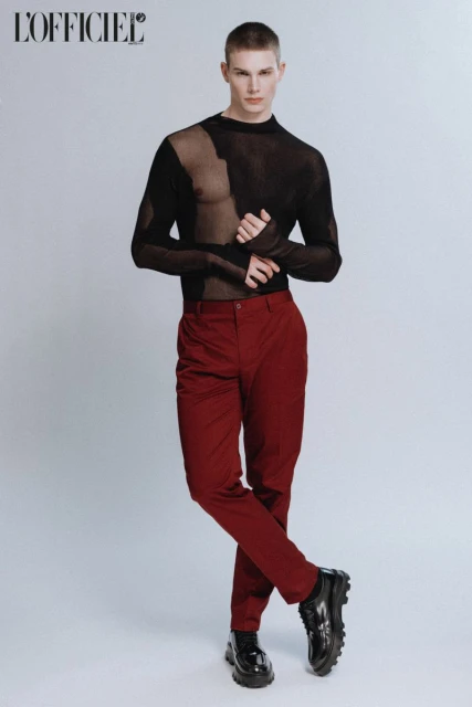 Male model posing in a studio wearing a black sheer top and burgundy pants, glossy black shoes, and a buzzcut hairstyle.