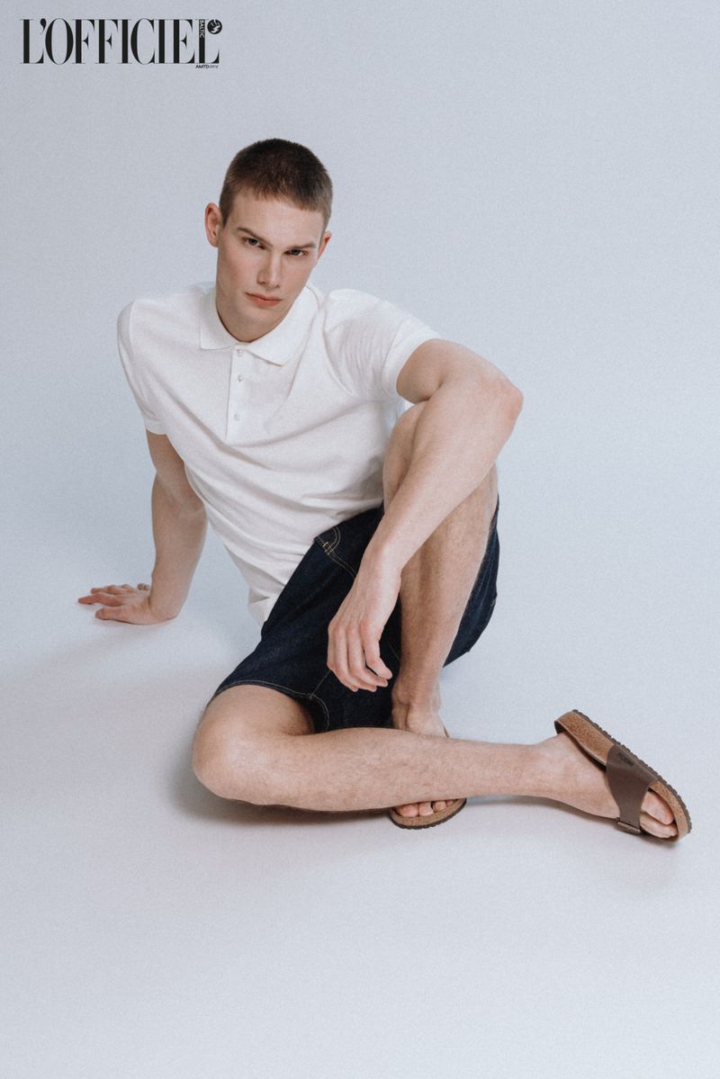 A fair-skinned young man sits on a white surface wearing a white polo shirt, dark shorts, and brown sandals in a studio setting.