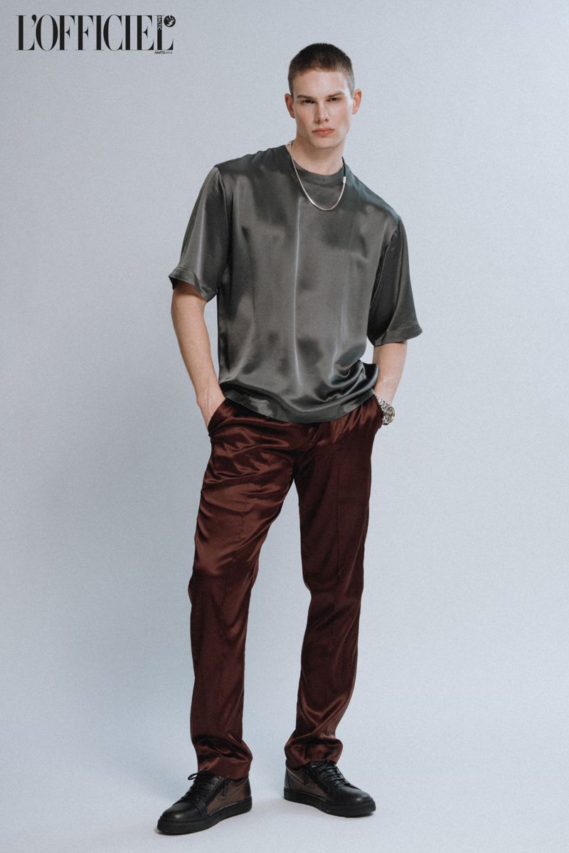 Full-length studio portrait of a fair-skinned adult male in satin top and trousers with a silver chain and black sneakers.