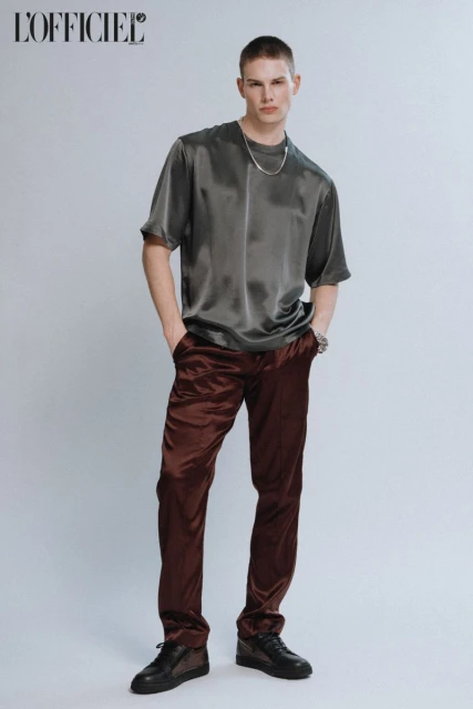 Full-length studio portrait of a fair-skinned adult male in satin top and trousers with a silver chain and black sneakers.